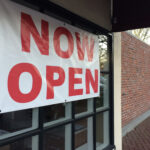 the new store hangging the red font on the white vinyl now open sign  the on the windows of store near the side walk